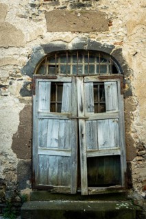 A rustic wooden door stands ajar in an aged stone wall, showing signs of weathering and decay. The