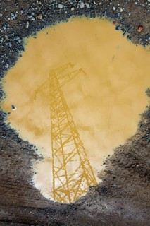 A power line tower is reflected in a large puddle of muddy water. The cloudy sky is visible, adding