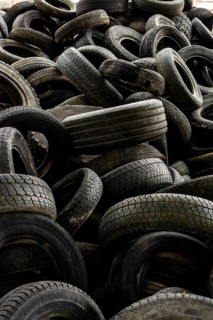 A large collection of worn tires is heaped together in a cluttered industrial setting. The scene