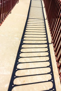 Bright sunlight creates intricate shadows from a red railing onto a stone pathway in a park,