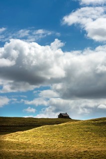 Ruined stone cottage stands alone on a grassy hillside under a partly cloudy sky in a remote