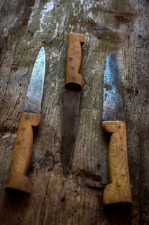 Three vintage kitchen knives are arranged on a weathered wooden surface. The blades display signs