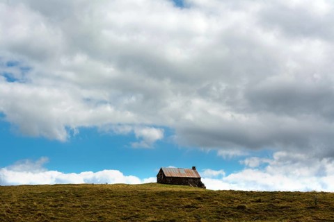 A small cabin sits alone atop a grassy hill, surrounded by wide-open space. The sky is filled with