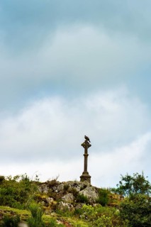 A stone cross towers over rocky ground, surrounded by greenery. Dark clouds loom overhead, creating
