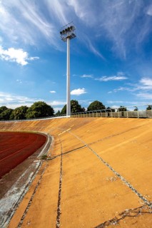 Bright daylight reveals an empty sports stadium with a vibrant red track. The tall floodlights,