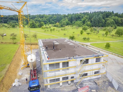 Large building shell with crane and workers in a rural setting, green roof, house construction,