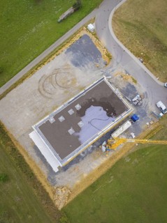 Aerial view of a rectangular building in shell construction on a large site next to a road, green