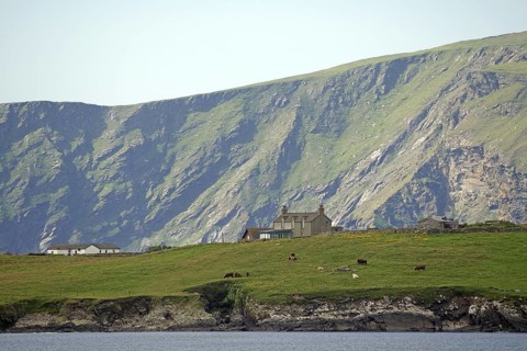 A remote house stands in green pasture against a dramatic mountain backdrop, Sumburgh, Shetland