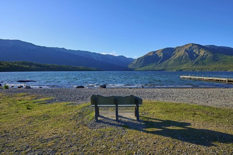 A park bench on the shore of a lake with a view of the surrounding mountains and a boardwalk under