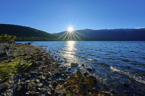 The sun rises above the horizon, pouring rays over the lake and the rocky shore, summer, Lake