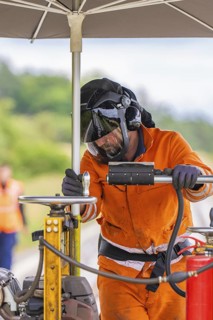 A worker in an orange suit and helmet works under an umbrella on a railway line, grinding work on