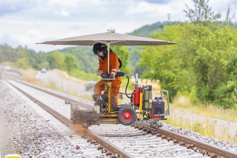 Worker in orange overalls operating a machine on railway tracks, protected by a large umbrella,