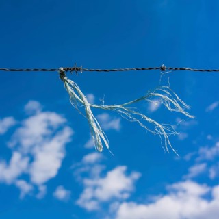 A frayed string hangs from a barbed wire fence, contrasting with the bright blue sky dotted with