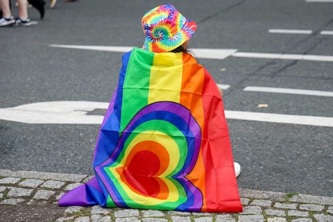 A participant with a rainbow flag and colourful hat sits on the side of the road at the 47th