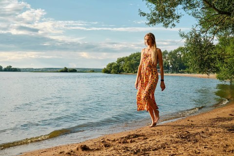 A woman in a colourful sundress strolls barefoot along the sandy shore of a tranquil lake. Lush