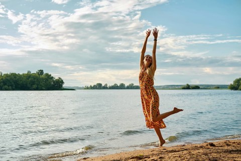 A carefree woman in a colourful sundress dances with joy along the riverbank on a sunny afternoon