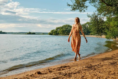 A woman in a vibrant sundress strolls barefoot along the sandy beach beside a calm lake. Sunshine