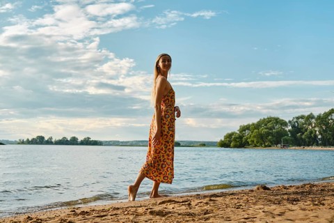 A woman is strolling barefoot along a sandy riverbank, wearing a colourful floral sundress. The