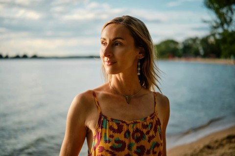 A woman stands near the water's edge, basking in the warm glow of sunset. She wears a colorful