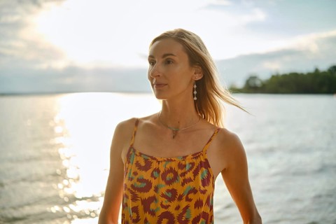 A woman with long hair poses by a calm body of water during sunset, wearing a vibrant patterned