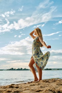 A woman stands gracefully on the sandy shore, her floral dress flowing in the wind. She gazes