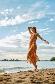 A woman stands barefoot on sandy shores, wearing a bright dress with a bold floral pattern. She
