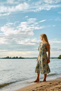 A woman wearing a floral dress stands barefoot on the sandy shore of a lake, gazing at the water.