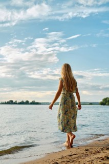 Rear view of barefoot woman standing on a sandy beach by a tranquil lake. The clear blue sky