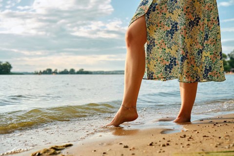 Unrecognizable woman stands at the edge of a lake, her bare foot touching the gentle waves at