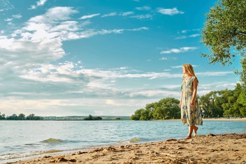 A woman in a floral dress strolls barefoot along the sandy shore of a peaceful lake, surrounded by