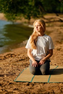 A woman sits on a mat by the riverside, engaged in a moment of mindfulness. She has long blonde