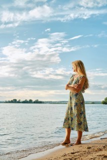 Thoughtful woman with long blonde hair stands on the sandy shore, gazing thoughtfully at the water