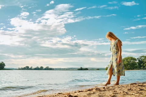 A woman wades along the sandy shore, wearing a colorful floral dress. The sun shines brightly, and