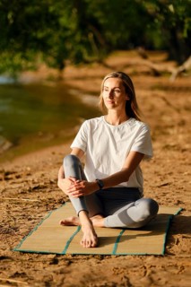 A woman sits cross-legged on a mat by a riverbank, focusing on mindfulness as the sun sets. She