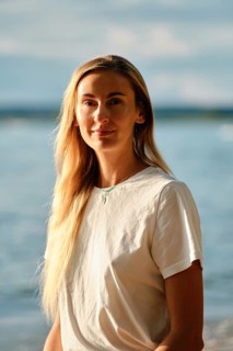 A woman with long hair smiles gently while standing near the water's edge at the beach. The bright