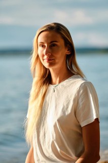 A woman with long blonde hair is standing near the water's edge at sunset. She gazes thoughtfully