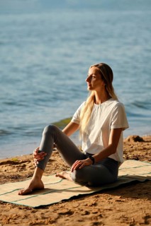A woman sits on yoga mat by the lakeshore and listens a surf noise. The calm water reflects the