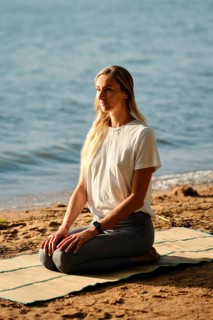 A woman sits calmly on a yoga mat by the water, practicing mindfulness as the sun sets. The serene