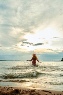 Funny woman plays joyfully in the water at a lakeside beach. The sun sets behind her, casting