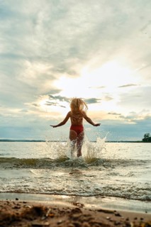A woman in red swimwear plays in the water, splashing joyfully as the sun sets in the background.