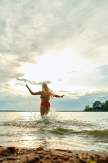A woman in a red swimsuit plays in the shallow water of a lake. The sun sets behind her, casting a