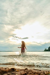 A woman with long hair playfully splashes in the water at a beach during sunset. The sky is