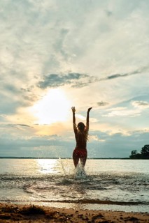 A woman stands in shallow water at a beach during sunset, joyfully raising her arms towards the
