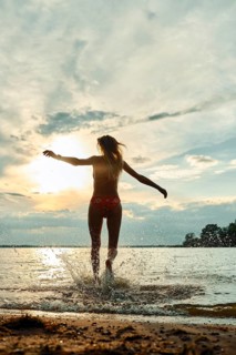 A woman wearing swimwear joyfully splashes through water at sunset. The sky displays vibrant