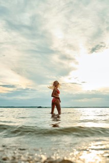 A woman wades through shallow water, smiling as she enjoys the warm sunset. The sky is painted with