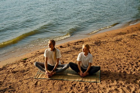 Two women are sitting on a mat by the water's edge and doing yoga poses as the sun begins to set.