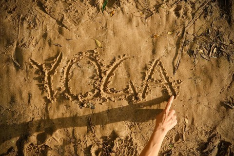 A person draws the word yoga in the sand using her finger. The setting is a sunny beach, showcasing