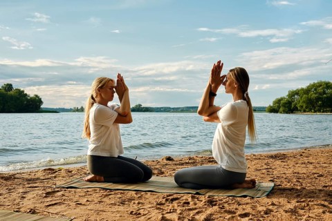 Two individuals are engaged in a yoga session on a sandy beach by a tranquil lake. The sun sets,