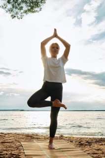 A woman performs yoga on a sandy beach, balancing in a tree pose with arms raised towards the sky.