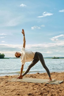 A person practices yoga on a mat by the water's edge during sunset. Woman stands in a triangle